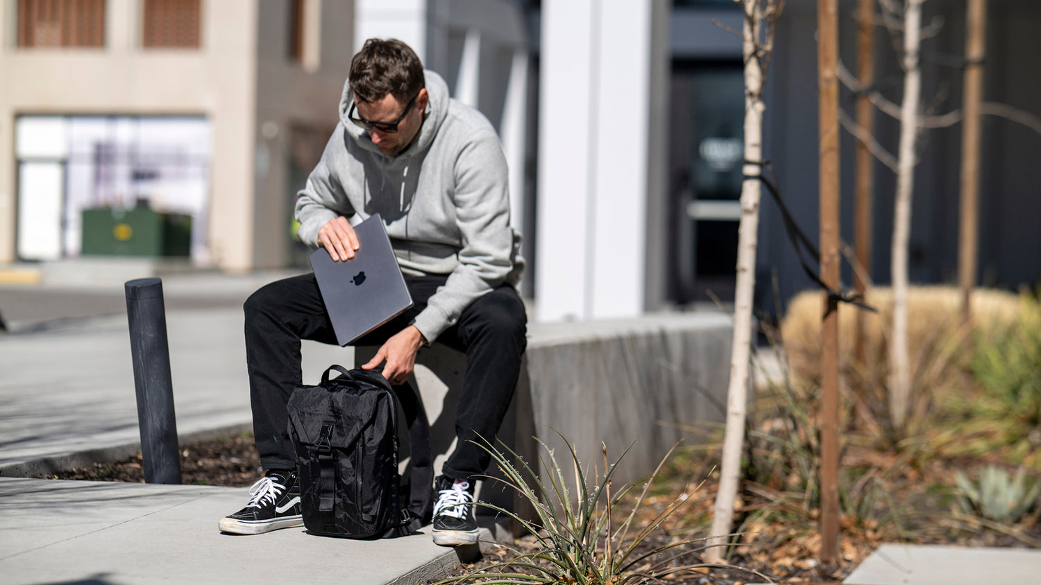 Person sitting on a sidewalk holding a laptop and a backpack